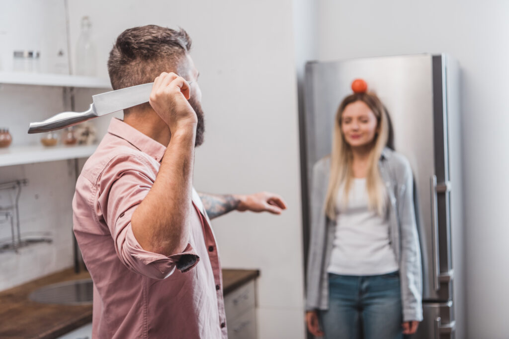 man throwing sharp knife at tomato on woman head